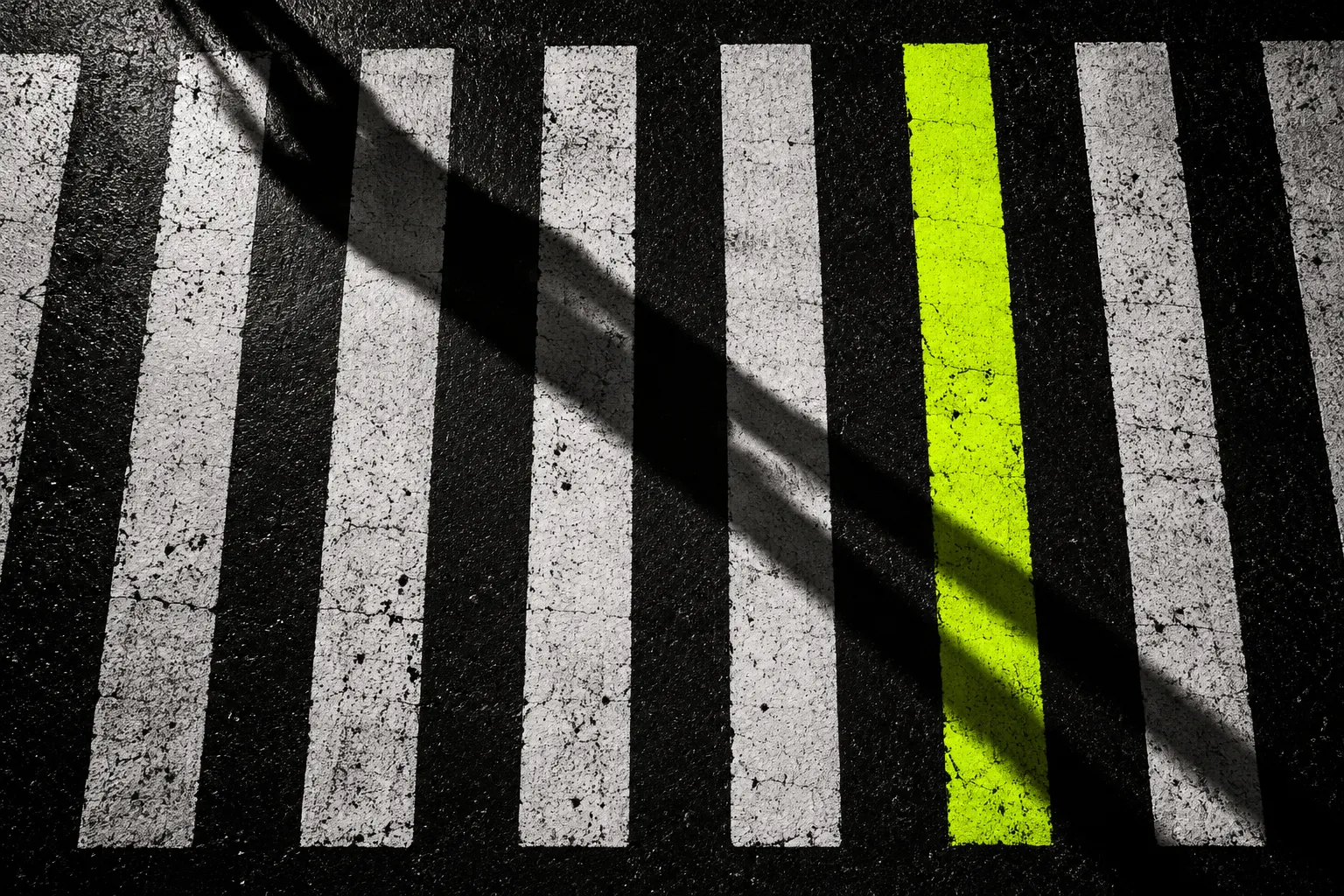 A pedestrian crossing at night from above, one long shadow crossing, one stripe yellow-green.